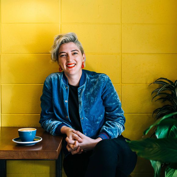 A photo of Beth Brash sitting against a bright yellow wall.